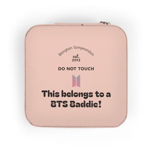 May include: A square, light pink jewelry travel case with a zipper closure. The top features the text Bangtan Sonyeondan est. 2013, DO NOT TOUCH, a BTS logo, and the text This belongs to a BTS Baddie! printed in black and a gradient pink color.