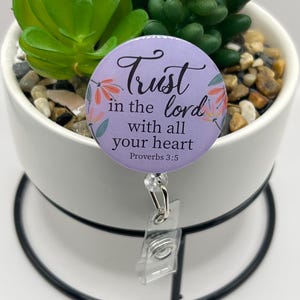 Puede incluir: Un carrete de insignia redondo, de color lavanda, con el texto "Trust in the Lord with all your heart" y detalles florales. El carrete de insignia está sujeto a un soporte de plástico transparente y a un clip metálico plateado. El carrete de insignia está delante de una pequeña planta en maceta.