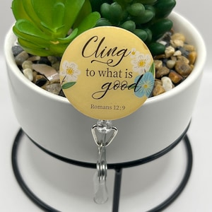 Puede incluir: Un portatarjetas amarillo con el texto "Cling to what is good" y el versículo bíblico Romanos 12:9. El portatarjetas tiene un diseño floral y está sujeto a un clip de plástico transparente. El portatarjetas está frente a una maceta blanca con una planta suculenta.