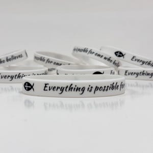 Puede incluir: Pulseras de goma blancas con texto negro y un símbolo de pez cristiano. Las pulseras tienen la frase "Todo es posible para el que cree". Las pulseras están apiladas y dispersas.