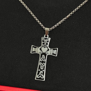 May include: Silver-toned pendant necklace featuring a Celtic cross design. The cross pendant has intricate details, including a heart, hands, and Celtic knot patterns. The necklace has a chain with a textured, woven appearance.