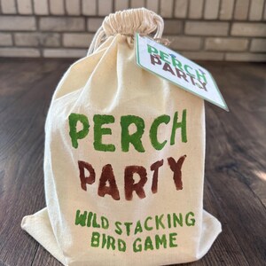 May include: A cream canvas bag with a drawstring top, printed with "PERCH PARTY" in green and brown, and "WILD STACKING BIRD GAME" in green. A small tag with "PERCH PARTY" is attached. The bag rests on a wooden surface.