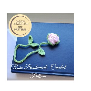 May include: A handmade crochet rose bookmark with a pink rose and green leaves. The bookmark is lying on a blue surface. The text "Rose Bookmark Crochet Pattern" is visible.