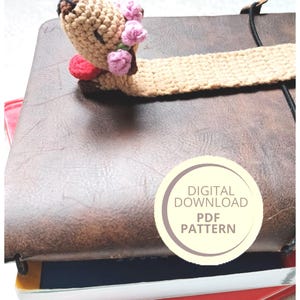 May include: A handmade crochet bookmark featuring a bear head with pink flowers and a red bow tie. The bookmark is attached to a brown leather journal. A digital PDF pattern is available.