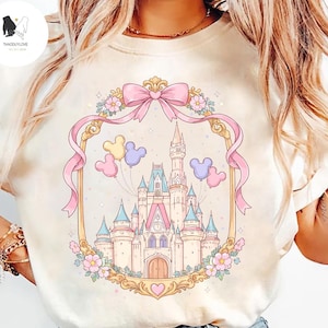 May include: A cream-coloured t-shirt with a fairytale castle illustration framed by a pink ribbon. The castle has blue and pink turrets, with Mickey Mouse balloons above. Floral details and the text "THADDUYLOVE" are also present in the design.