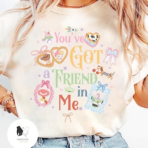 May include: Cream-colored sweatshirt featuring the phrase "You've Got a Friend in Me" in colorful, whimsical lettering. The design includes various cartoon character illustrations within decorative frames and star accents.