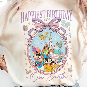 May include: Cream-colored t-shirt featuring a colorful graphic with Disney characters, a castle, and the text "Happiest Birthday On Earth." The design includes Mickey Mouse, Minnie Mouse, Donald Duck, and other characters. The graphic is framed by a light purple bow and decorative elements.