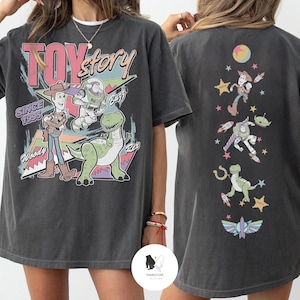 May include: Dark gray t-shirt featuring the words "Toy Story" with Woody, Buzz Lightyear, and Rex the dinosaur. The back of the shirt has a vertical design with various Toy Story characters and a colorful moon and stars.