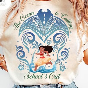 Op de afbeelding: Een cr&egrave;mekleurig T-shirt met een afbeelding van een stripfiguur die op een strand met palmbomen en oceaangolven rent. Het ontwerp bevat blauwe tribale patronen en de groene tekst "The Ocean is Calling" en "School's Out" in een sierlijk lettertype.