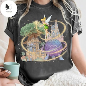 Op de afbeelding: Donkergrijze t-shirt met een kleurrijke afbeelding van Tinkerbell met een toverstaf, omringd door iconische Disney-park bezienswaardigheden zoals het Hollywood Tower Hotel, Spaceship Earth en Cinderella Castle. Het ontwerp is omlijnd met gouden glitters.
