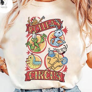 May include: Cream-colored t-shirt featuring a colorful graphic of circus characters. The design includes the text "P.T. Flea's Circus" in a red banner, with various insect illustrations in circular frames. The shirt has a crew neck and short sleeves.