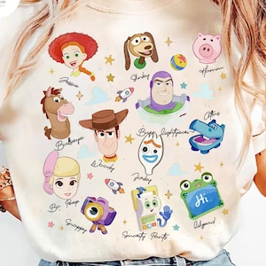 May include: Cream-colored t-shirt featuring colorful cartoon illustrations of characters from the movie Toy Story. The design includes images of Woody, Buzz Lightyear, Jessie, and others, with their names written below.
