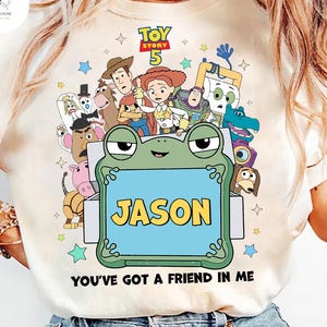May include: Cream-colored t-shirt featuring a colorful graphic of characters from Toy Story 5. The design includes a frog holding a sign that says "JASON" and the phrase "YOU'VE GOT A FRIEND IN ME".