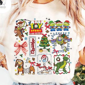 May include: Cream-colored t-shirt featuring a colorful Christmas-themed Toy Story graphic. The design includes characters like Woody, Buzz Lightyear, and the Little Green Men, along with a Christmas tree and the word "Christmas".