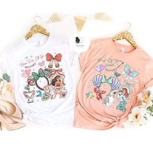 May include: Two white and peach-colored t-shirts with Disney-themed graphics. The white shirt features Moana and the text "How Far I'll Go." The peach shirt displays Ariel and the text "Part of Your World." Both shirts have gold hangers.