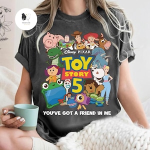 May include: Charcoal grey t-shirt with a vibrant graphic of characters from Disney Pixar's Toy Story 5. The design includes Woody, Buzz Lightyear, and other characters. The text "TOY STORY 5" and "YOU'VE GOT A FRIEND IN ME" are also displayed.
