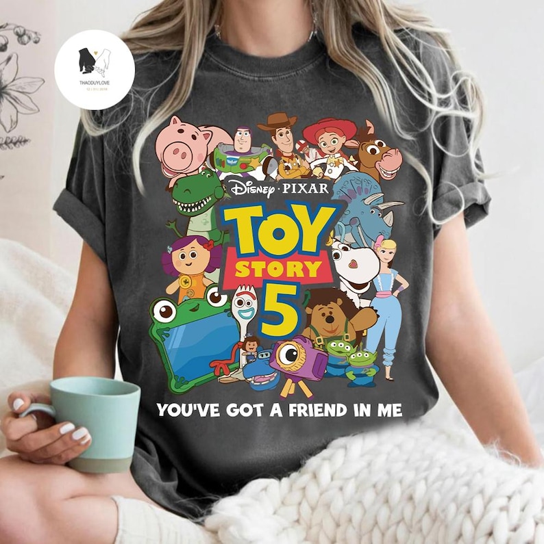 May include: Charcoal grey t-shirt with a vibrant graphic of characters from Disney Pixar's Toy Story 5. The design includes Woody, Buzz Lightyear, and other characters. The text "TOY STORY 5" and "YOU'VE GOT A FRIEND IN ME" are also displayed.