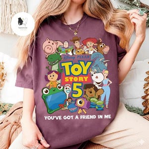 May include: A maroon t-shirt with a vibrant graphic of characters from the Disney Pixar film Toy Story. The design includes the text "Toy Story 5" and "You've Got a Friend in Me".