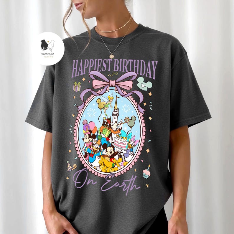 May include: Dark gray t-shirt with a colorful graphic that says "Happiest Birthday On Earth." The graphic features Disney characters, a castle, balloons, and a birthday cake. The design is framed by a pink and purple oval with a bow.