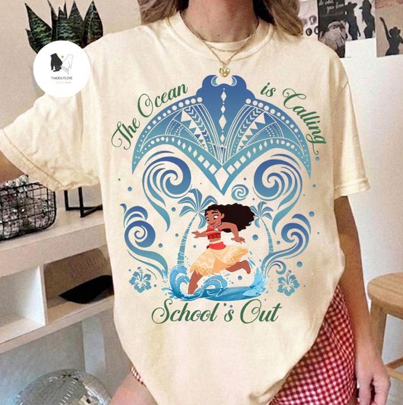 Op de afbeelding: Een cr&egrave;mekleurig t-shirt met korte mouwen en een afbeelding van een stripfiguur die op het water rent. Het ontwerp bevat blauwe golfpatronen, palmbomen en de tekst The Ocean is Calling en School's Out in groene letters.