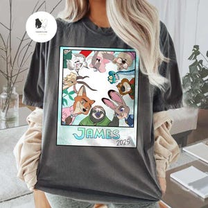 以下が含まれることがあります： ズートピアのキャラクターのカラフルな漫画のイメージが特徴の濃い灰色のTシャツ。画像は水色の枠で囲まれ、「JAMES」という名前と「2025」という年が含まれています。