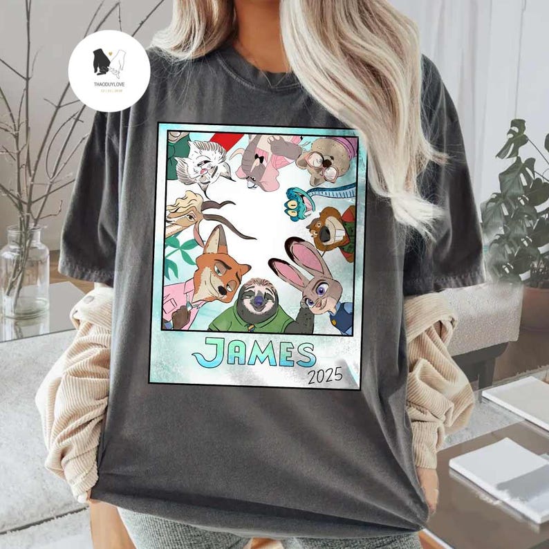 以下が含まれることがあります： ズートピアのキャラクターのカラフルな漫画のイメージが特徴の濃い灰色のTシャツ。画像は水色の枠で囲まれ、「JAMES」という名前と「2025」という年が含まれています。