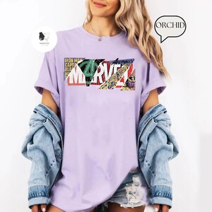 Op de afbeelding: Een lichtpaars T-shirt met het Marvel-logo in een collage-stijl. Het logo bevat stripboekafbeeldingen en het woord "MARVEL" in het wit. Het shirt heeft korte mouwen en een losse pasvorm.