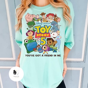 May include: A light teal t-shirt with a colourful graphic of characters from the Disney Pixar film Toy Story 5. The graphic includes the film title and the phrase "You've Got a Friend in Me".