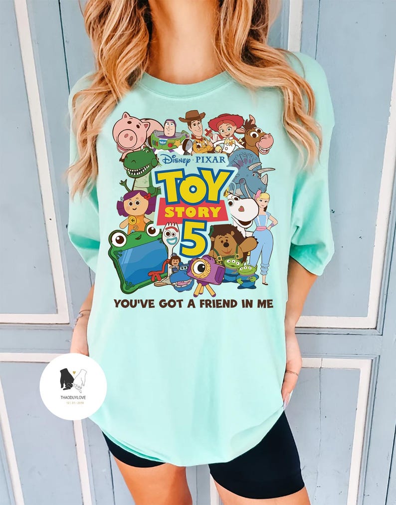 May include: A light teal t-shirt with a colourful graphic of characters from the Disney Pixar film Toy Story 5. The graphic includes the film title and the phrase "You've Got a Friend in Me".