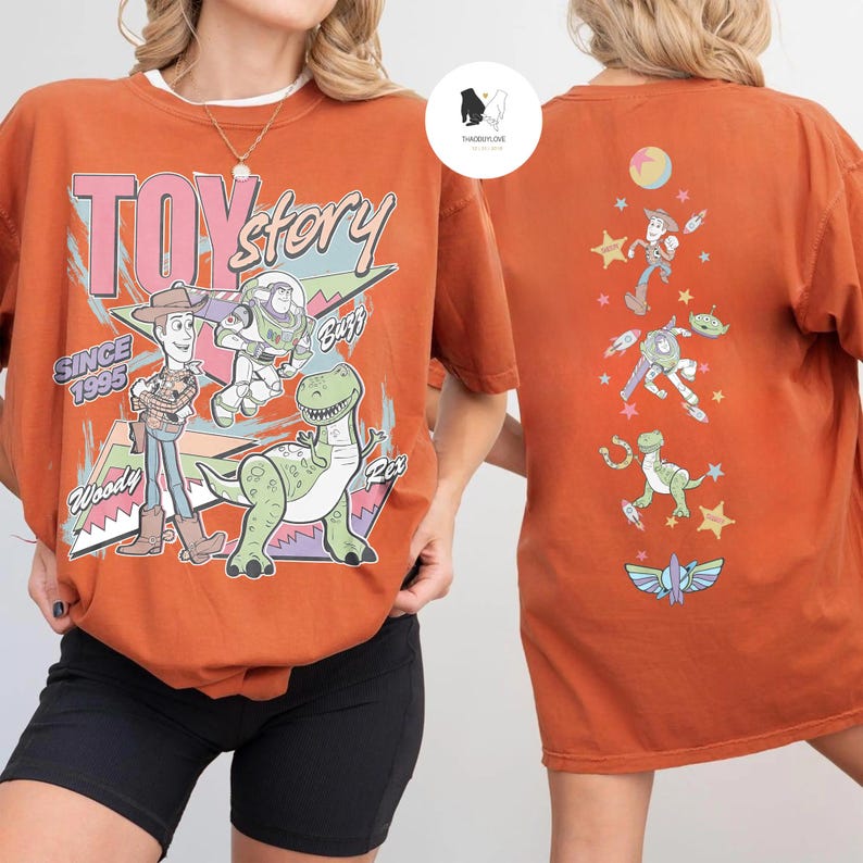 May include: Burnt orange t-shirt featuring the Toy Story logo and characters Woody, Buzz, and Rex. The front of the shirt displays the text "TOY STORY" and "SINCE 1995". The back of the shirt has a vertical design with more Toy Story characters.