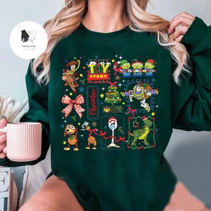 May include: Dark green sweatshirt featuring a colorful Toy Story Christmas design. The graphic includes characters like Woody, Buzz Lightyear, and the Little Green Men, along with a Christmas tree and bow. The words "Toy Story" and "Christmas" are also featured.