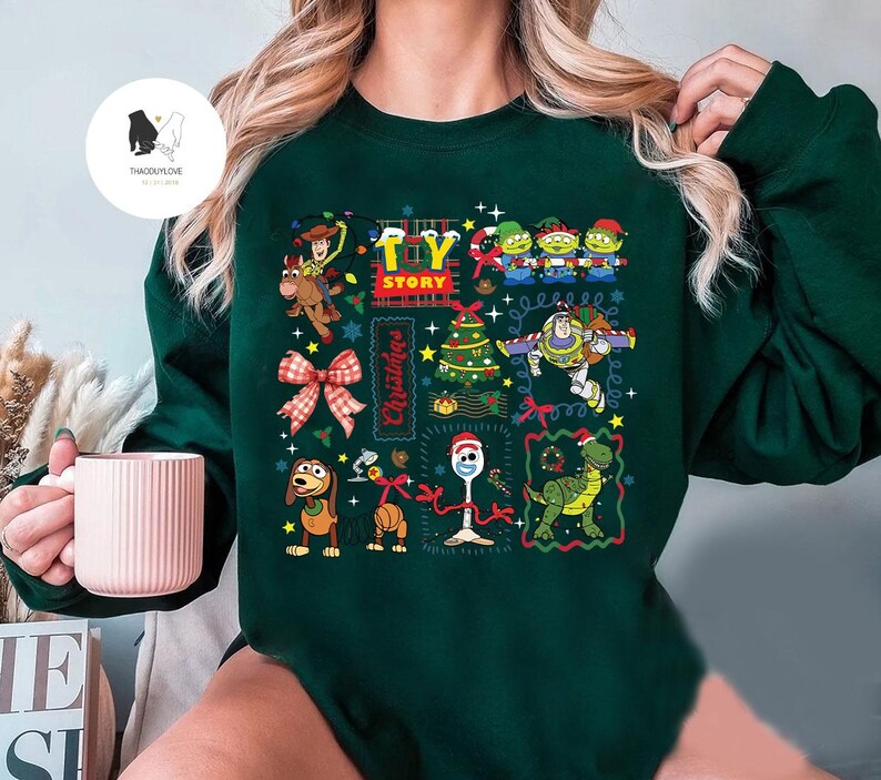 May include: Dark green sweatshirt featuring a colorful Toy Story Christmas design. The graphic includes characters like Woody, Buzz Lightyear, and the Little Green Men, along with a Christmas tree and bow. The words "Toy Story" and "Christmas" are also featured.