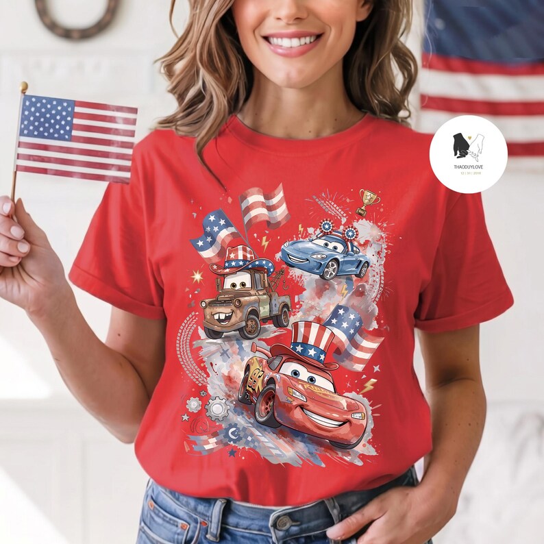 Op de afbeelding: Een felrood T-shirt met korte mouwen en een grafische print van twee cartoonauto's, een rode en een bruine, die patriottische hoeden dragen. Het ontwerp bevat Amerikaanse vlagmotieven, sterren en een gouden trofee op een gespatte achtergrond.