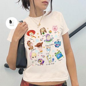 May include: Cream-colored t-shirt featuring colorful cartoon character illustrations. The shirt showcases characters like Woody, Buzz Lightyear, and Jessie, with their names written below. A fun and playful design.