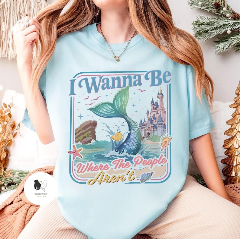 May include: Light blue t-shirt with a mermaid graphic. The graphic includes a mermaid tail, castle, and the text "I Wanna Be Where The People Aren't". The design features a mermaid tail, seashells, and a starfish.