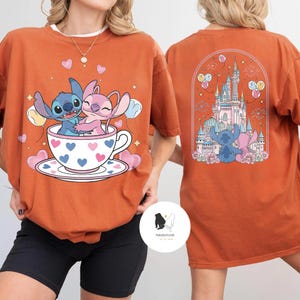 May include: Burnt orange t-shirt featuring a cartoon illustration of Stitch and Angel in a teacup with hearts. The back of the shirt shows a castle scene with Stitch and Angel. The shirt has a crew neck and short sleeves.