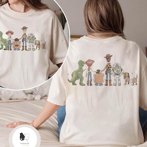 Peut inclure: T-shirt cr&egrave;me avec une illustration de personnages de dessins anim&eacute;s populaires. Le design comprend un dinosaure vert, une cowgirl, une t&ecirc;te de pomme de terre, un cow-boy, un ranger de l'espace et un chien. Le dos du t-shirt a &eacute;galement le m&ecirc;me design.