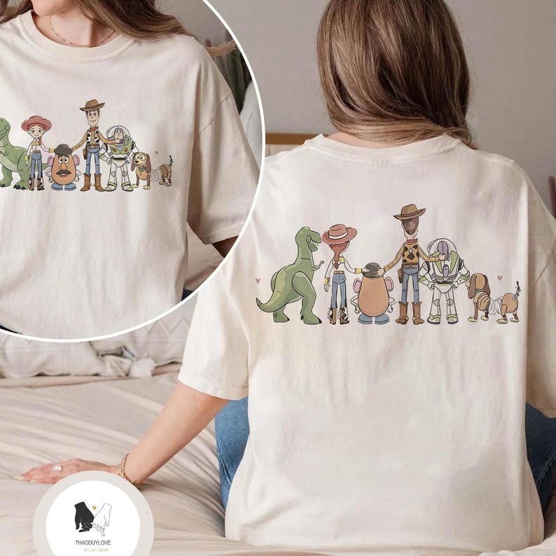Peut inclure: T-shirt cr&egrave;me avec une illustration de personnages de dessins anim&eacute;s populaires. Le design comprend un dinosaure vert, une cowgirl, une t&ecirc;te de pomme de terre, un cow-boy, un ranger de l'espace et un chien. Le dos du t-shirt a &eacute;galement le m&ecirc;me design.