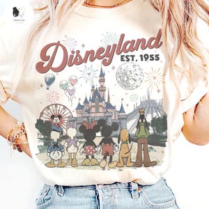 May include: A cream-coloured t-shirt featuring a graphic print of Disneyland with the text "Disneyland EST. 1955". The design includes a castle, a ferris wheel, fireworks, a moon, and cartoon characters including Mickey, Minnie, Donald, Daisy, Goofy, and Pluto.