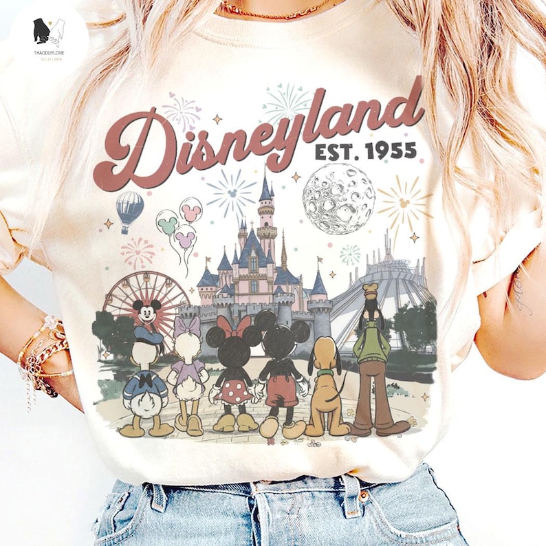 May include: A cream-coloured t-shirt featuring a graphic print of Disneyland with the text "Disneyland EST. 1955". The design includes a castle, a ferris wheel, fireworks, a moon, and cartoon characters including Mickey, Minnie, Donald, Daisy, Goofy, and Pluto.
