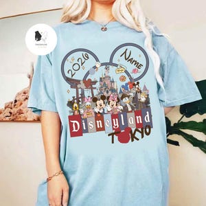 May include: Light blue t-shirt featuring a Disneyland Tokyo design. The design includes Mickey Mouse ears with the year 2026 and a space for a name. The graphic showcases Disney characters and a castle, with the words "Disneyland Tokyo" in a stylized font.