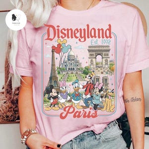 May include: Pink t-shirt featuring a Disneyland Paris graphic. The design includes the Eiffel Tower, Arc de Triomphe, and cartoon characters Mickey Mouse, Minnie Mouse, Donald Duck, Daisy Duck, Goofy, and Pluto. Text reads "Disneyland Paris Est. 1992".