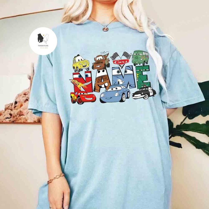 K&ouml;nnte beinhalten: Hellblaues T-Shirt mit dem Wort "NAME", das aus Cartoon-Autos besteht. Das Design zeigt Charaktere aus dem Film Cars, darunter Lightning McQueen und Mater. Das Shirt ist ein kurz&auml;rmeliger, l&auml;ssiger Stil.