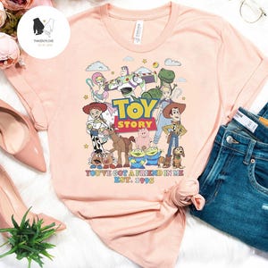 May include: A pink t-shirt with a graphic of the Toy Story characters, including Woody, Buzz Lightyear, Jessie, and Rex. The text "Toy Story" and "You've got a friend in me Est. 1995" is printed on the shirt.