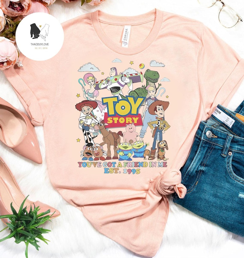 May include: A pink t-shirt with a graphic of the Toy Story characters, including Woody, Buzz Lightyear, Jessie, and Rex. The text "Toy Story" and "You've got a friend in me Est. 1995" is printed on the shirt.
