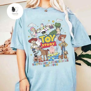 May include: A light blue t-shirt with a graphic of the Toy Story characters. The characters are arranged in a circle around the words "Toy Story". The text "You've got a friend in me. Est. 1995" is printed below the characters.