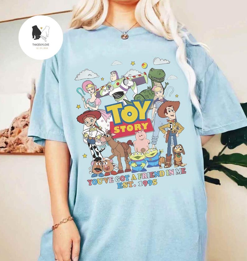May include: A light blue t-shirt with a graphic of the Toy Story characters. The characters are arranged in a circle around the words "Toy Story". The text "You've got a friend in me. Est. 1995" is printed below the characters.