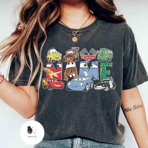 K&ouml;nnte beinhalten: Dunkelgraues T-Shirt mit dem Wort "Cars", das mit Zeichentrick-Autofiguren geschrieben ist. Das Design umfasst ein gelbes Auto, einen braunen LKW, ein rotes Rennauto und ein blaues Auto. Das Shirt hat einen Rundhalsausschnitt und kurze &Auml;rmel.