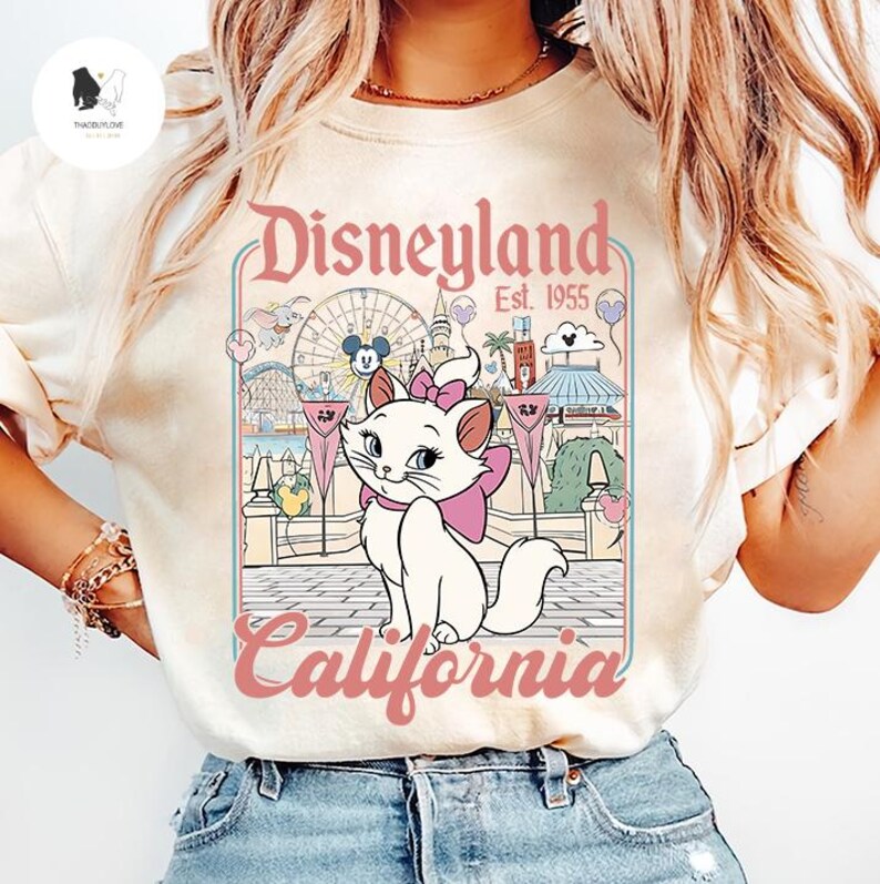 Peut inclure: T-shirt cr&egrave;me avec un motif Disneyland. Le design comprend le texte "Disneyland California Est. 1955" et une illustration de Marie, la chatte blanche des Aristochats, avec un n&oelig;ud rose. L'arri&egrave;re-plan repr&eacute;sente des attractions du parc.