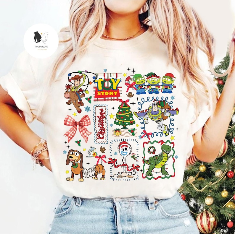 May include: Cream-colored t-shirt featuring a colorful Christmas-themed Toy Story graphic. The design includes characters like Woody, Buzz Lightyear, and the Little Green Men, along with a Christmas tree and the word "Christmas".